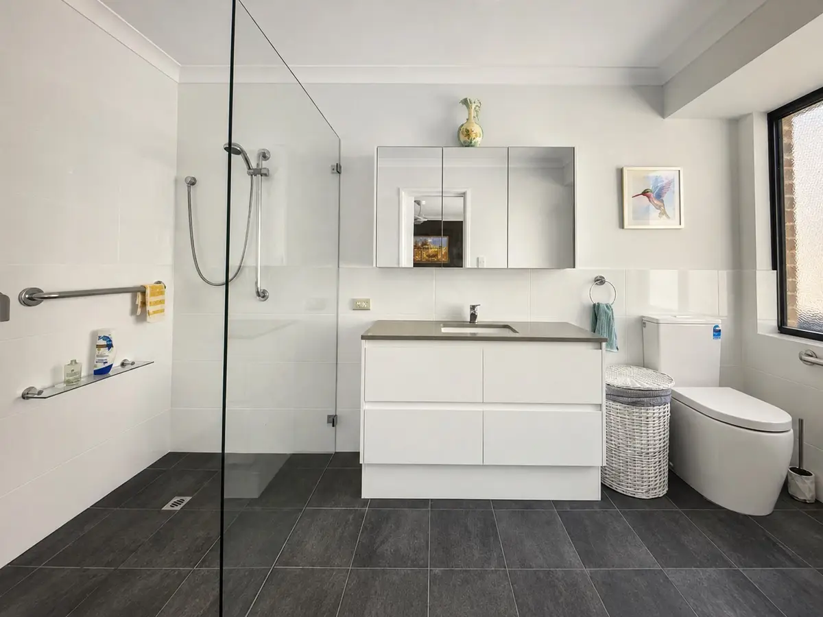 After: the Hocking ensuite with an open shower behind a single glass panel, a hand shower on a rail, grab rails, a drawer vanity, a cabinet mirror and dark non-slip floor tiles
