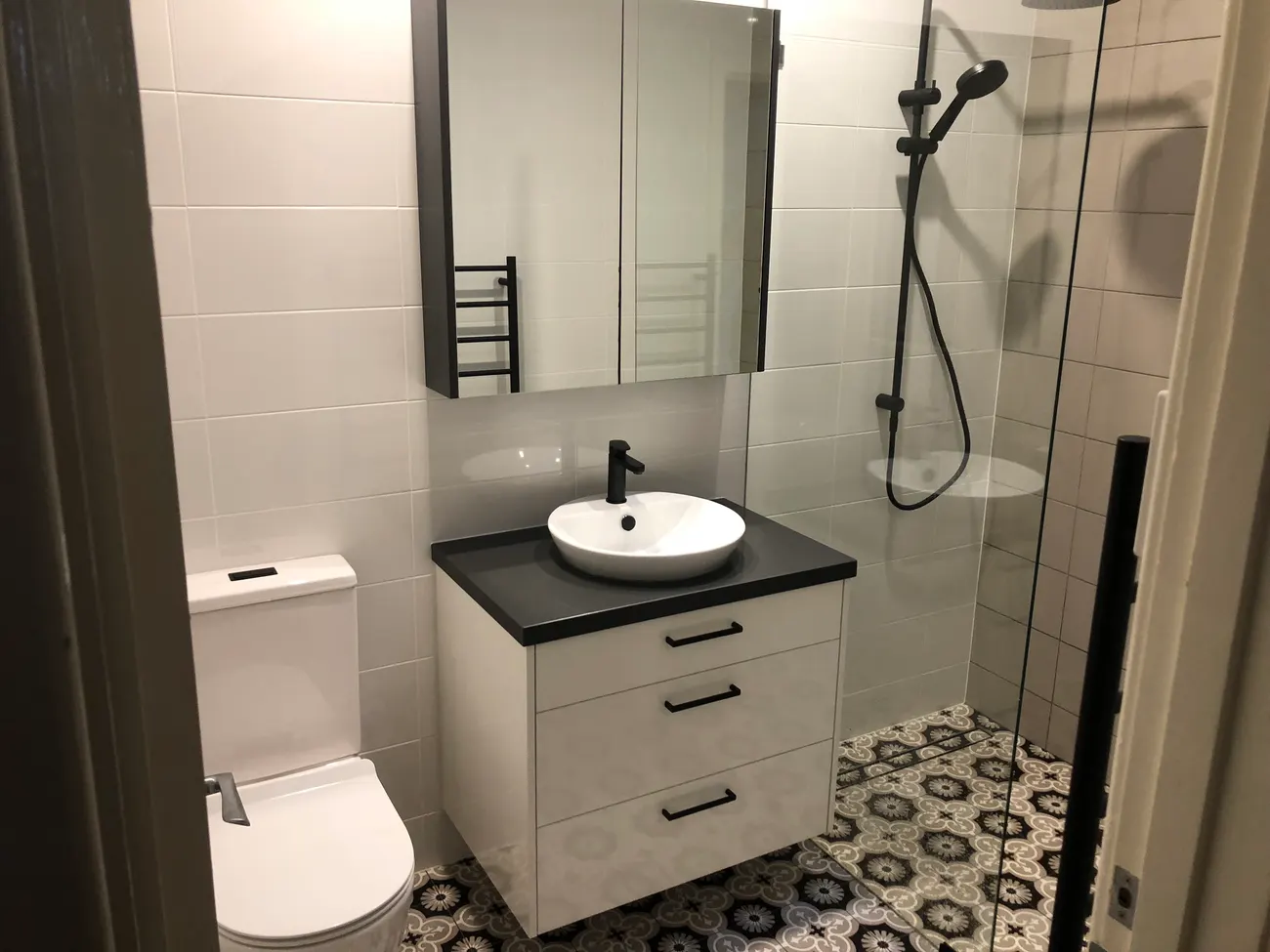 After: the same bathroom with a level walk-in shower behind frameless glass, patterned floor tiles, a wall-hung vanity and black tapware