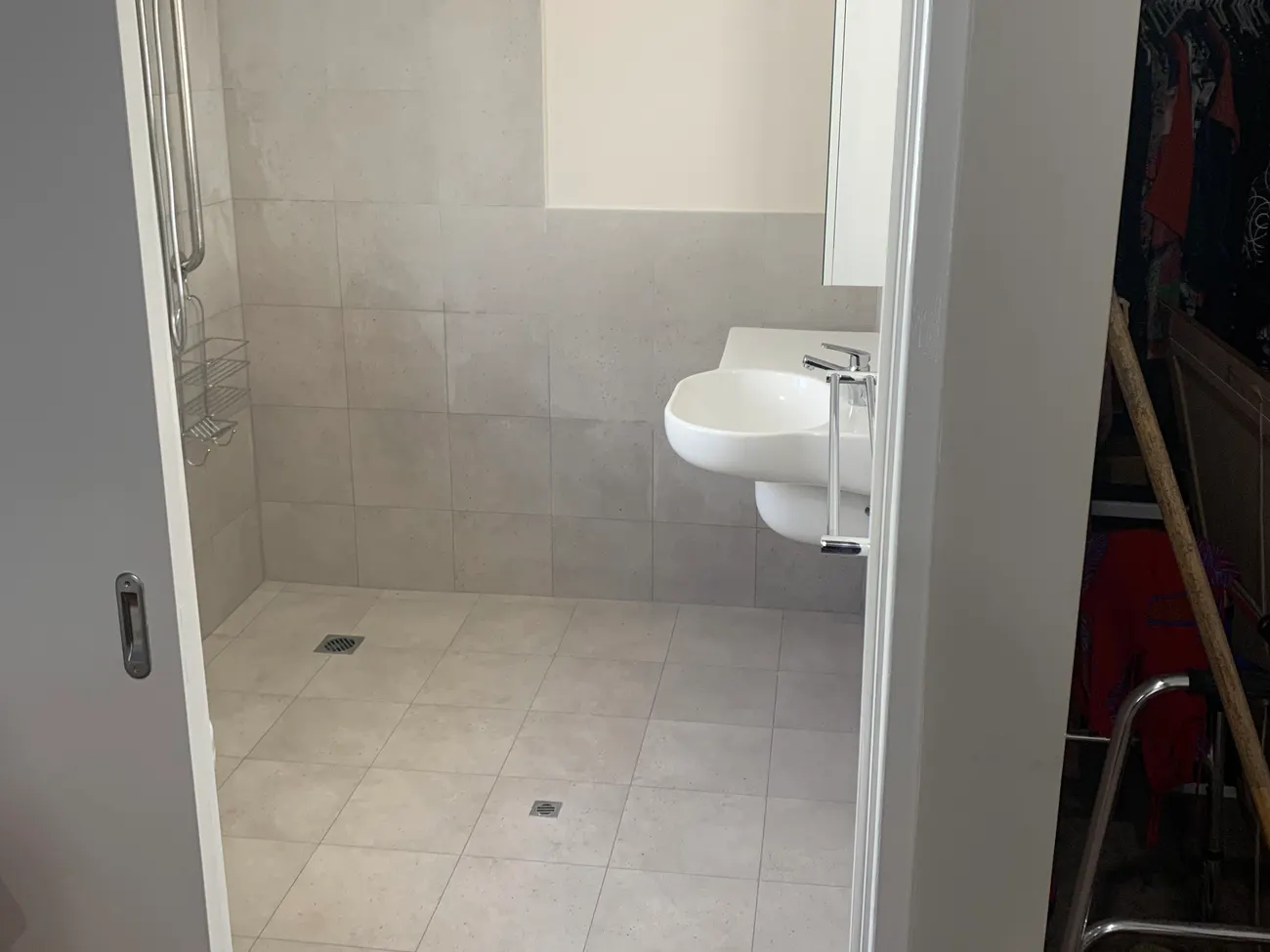 After: the same room as a wet room, with a completely level tiled floor draining to two floor wastes, a hand shower on a grab rail and a wall-hung basin
