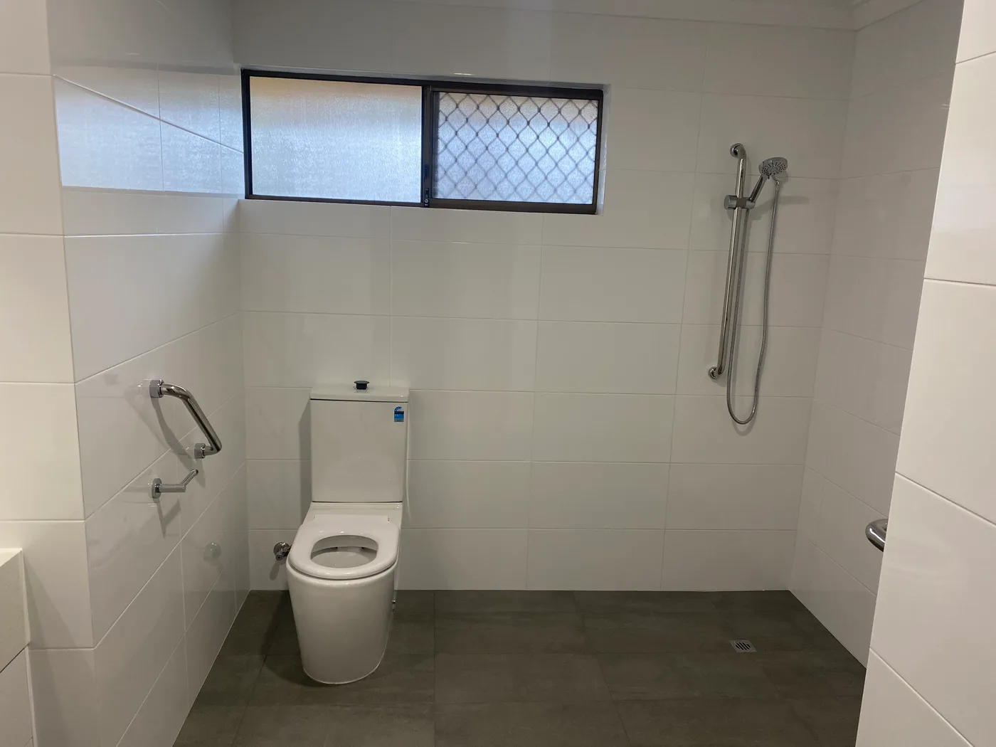 Open wet room in a Perth home with a completely level tiled floor, a floor waste, a hand shower on a rail and grab rails either side of the toilet